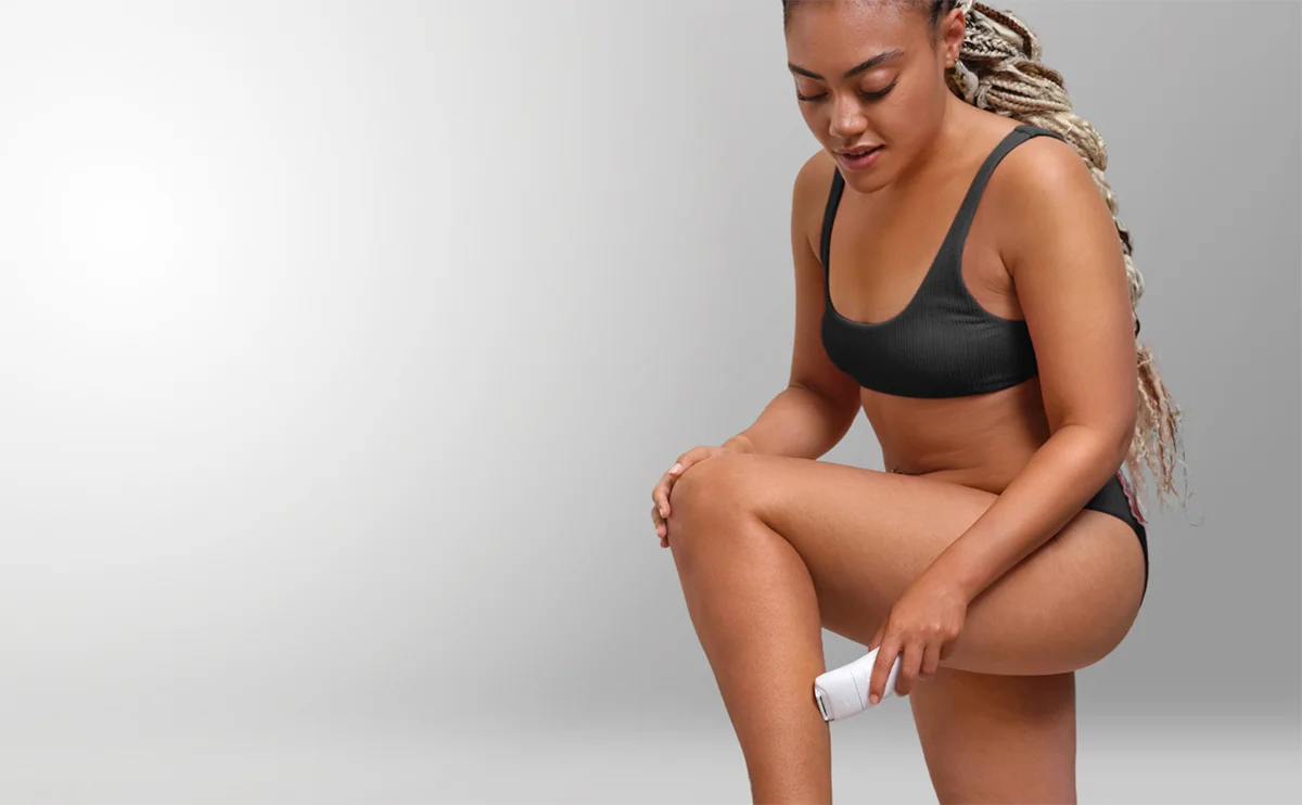 Kobieta w ciemnym bikini, z warkoczami, depilująca lewą nogę białym depilatorem na neutralnym tle.