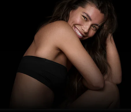 Uśmiechnięta kobieta w czarnym topie siedzi bokiem i patrzy w obiektyw na ciemnym tle.