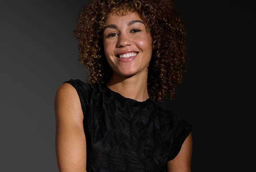 Uśmiechnięta kobieta z kręconymi włosami patrzy w obiektyw na ciemnym tle.