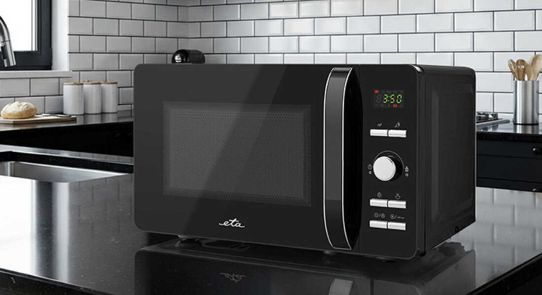 Kuchenka mikrofalowa ETA Gavento ETA120990010 Zdjęcie ukazuje urządzenie stojące na czarnym, połyskującym blacie w kuchni z białymi płytkami i czarną armaturą. Czarno‑srebrny front mikrofalówki kontrastuje z jasnym otoczeniem