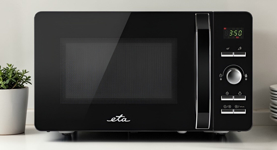 Kuchenka mikrofalowa ETA Gavento ETA120990010 Zdjęcie przedstawia tę samą kuchenkę ustawioną na jasnym blacie obok rośliny w doniczce, z czarnym frontem odbijającym światło. Front i panel sterowania są wyraźnie wyeksponowane równomierne grzanie brak zimnych miejsc wyświetlacz cyfrowy kontrola czasu timer 60 minut