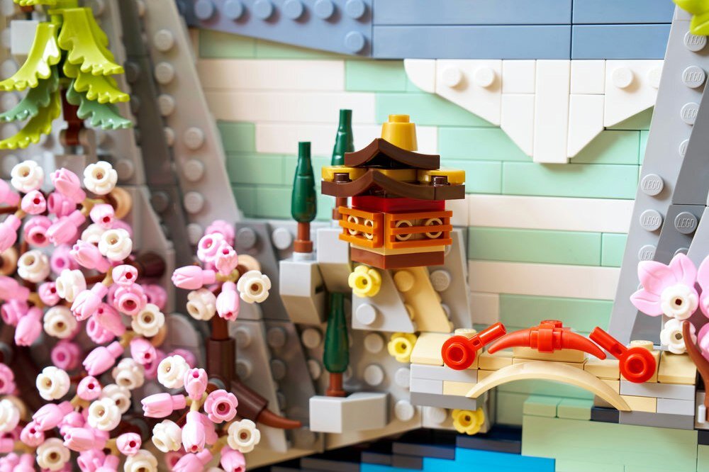 Na obrazie widać konstrukcję z klocków LEGO przedstawiającą japoński krajobraz z kwitnącą wiśnią, małym mostkiem i pagodą. Klocki są w różnych kolorach, w tym różowym, zielonym i szarym.