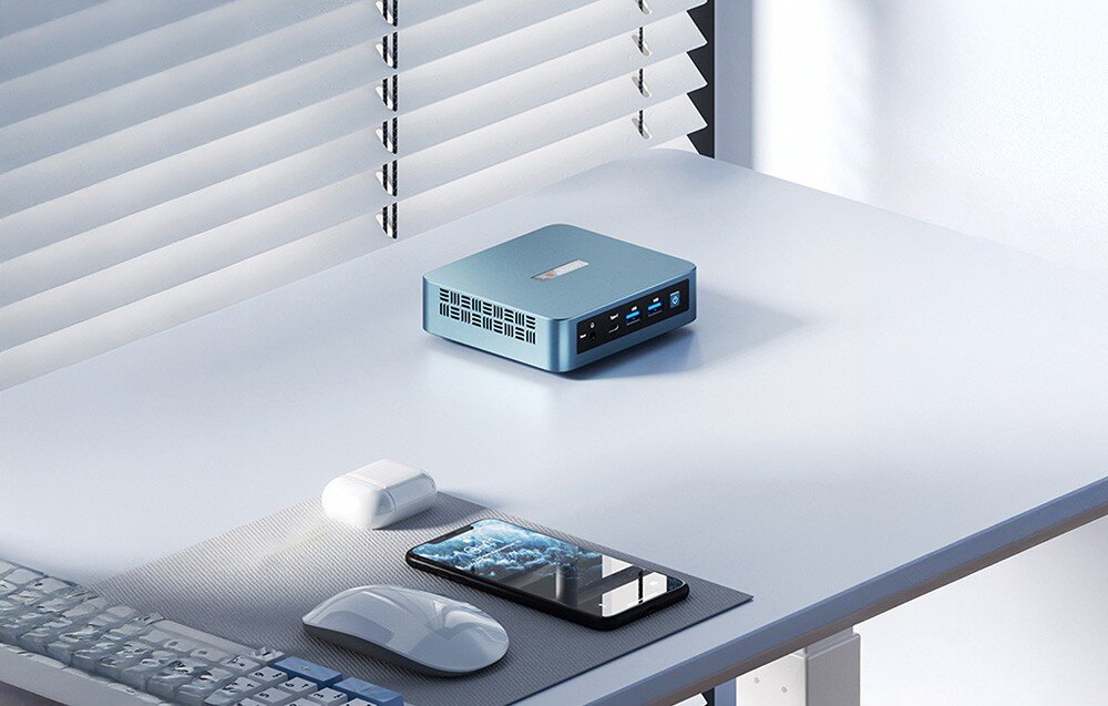 Komputer PELADN WI-6 N95 Urządzenie typu mini PC leżące na jasnym blacie obok smartfona, myszki komputerowej i słuchawek bezprzewodowych przy oknie z żaluzjami