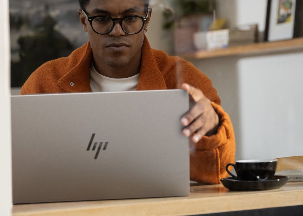 Mężczyzna w pomarańczowej kurtce pracujący na laptopie HP OmniBook w srebrnej obudowie, siedzący przy drewnianym stole w kawiarni z filiżanką kawy, skupiony na ekranie, eleganckie środowisko pracy zdalnej. - procesor Intel Core Ultra   