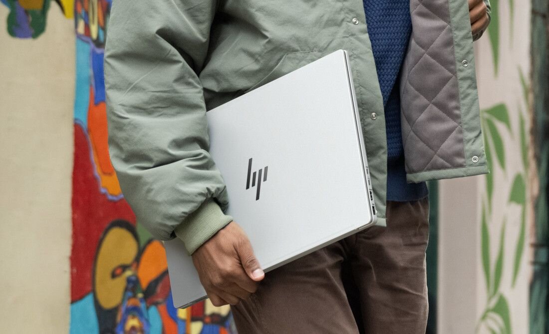 Mężczyzna w zielonej kurtce trzymający zamknięty laptop HP OmniBook w srebrnej obudowie na tle kolorowego muralu, podkreślający mobilność i łączność Wi-Fi 6E w miejskim otoczeniu. - Wi-Fi 6E Bluetooth 5.3  