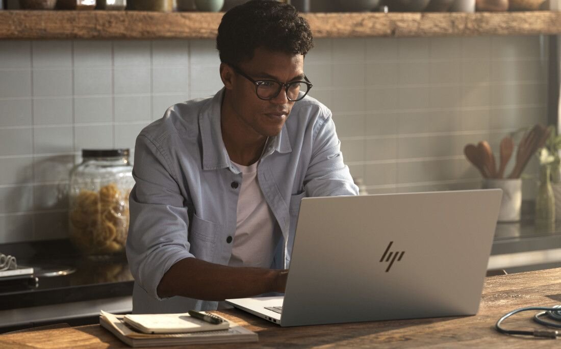 Młody mężczyzna w okularach pracujący na laptopie HP OmniBook w kuchni, podkreślający bezpieczeństwo danych dzięki modułowi TPM i komfort pracy zdalnej. - Fizyczna osłona kamery, Trusted Platform Module 