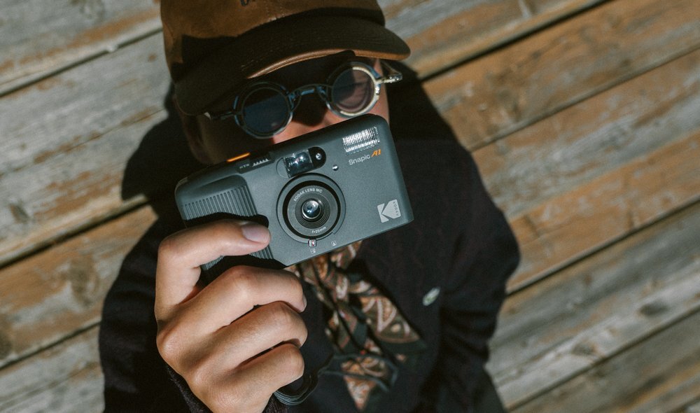 Osoba trzyma szary aparat analogowy z widocznym napisem \'KODAK Snapic A1\'. W tle znajduje się drewniana powierzchnia.