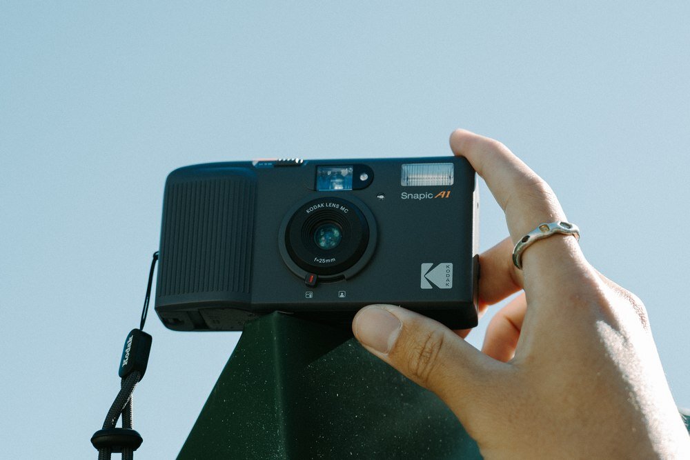Osoba trzyma szary aparat analogowy z napisem \'Snapic A1\' i logo \'KODAK\'. Aparat ma widoczny obiektyw i lampę błyskową.