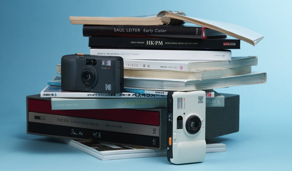 Na stosie książek znajdują się dwa aparaty analogowe, jeden czarny i jeden biały. Na czarnym aparacie widoczne jest logo \'KODAK\'.