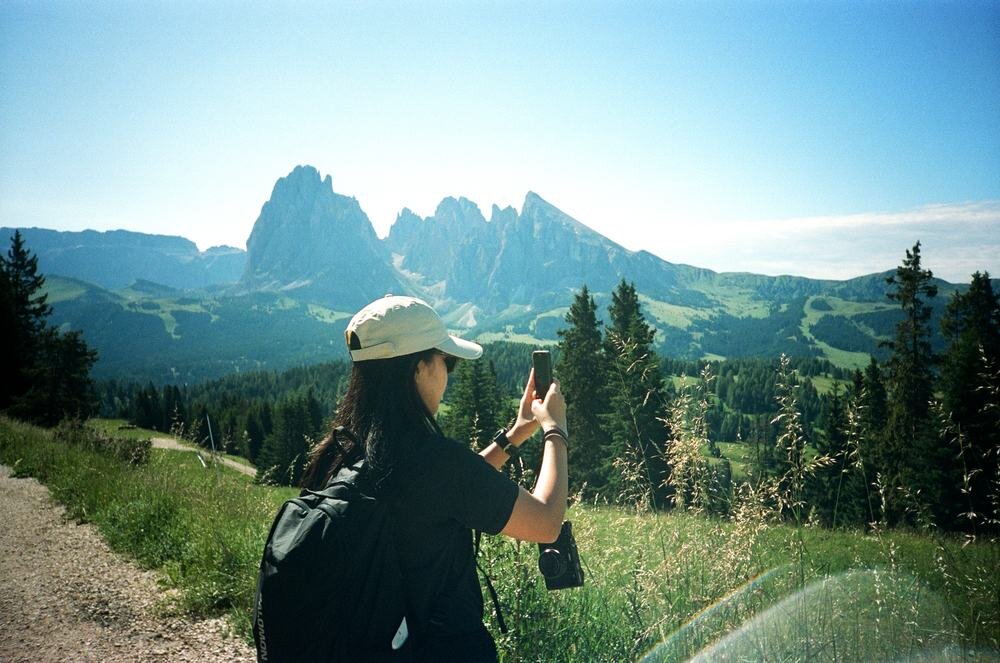 Osoba w czapce z daszkiem robi zdjęcie górskiego krajobrazu za pomocą smartfona. W tle widoczne są wysokie góry i zielone łąki.