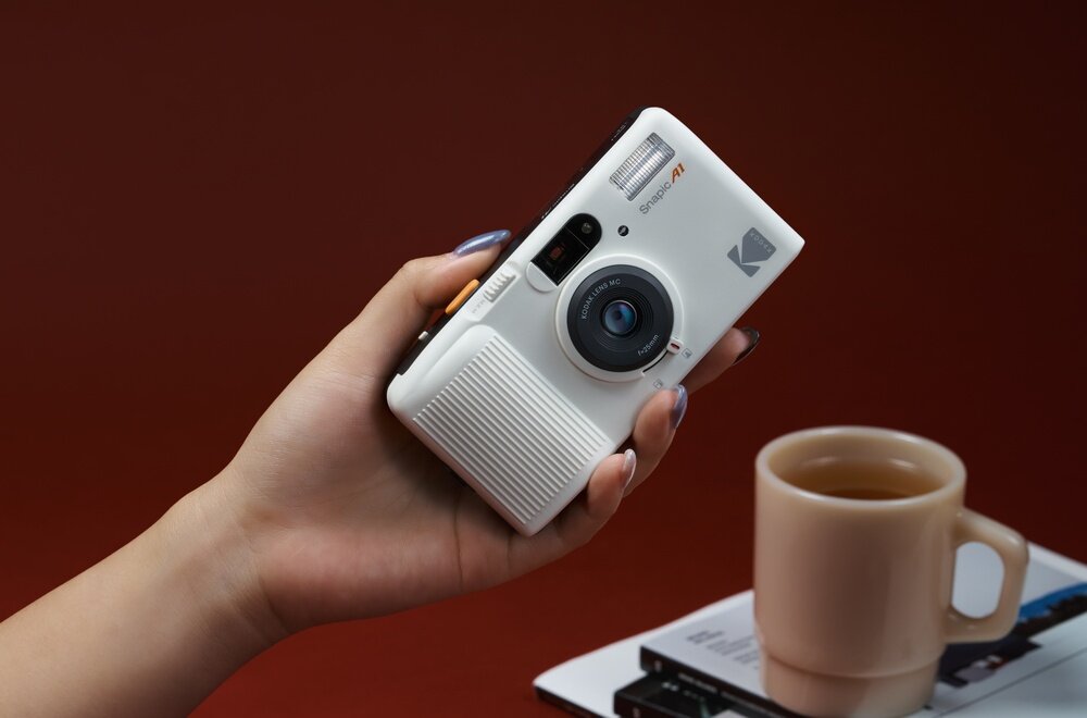 Osoba trzyma biały aparat analogowy z logo KODAK, obok stoi filiżanka z herbatą na książce. Na aparacie widoczny jest napis \'Snapic A1\'.