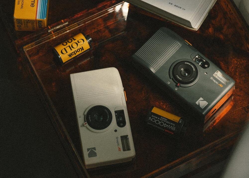 Na drewnianym stole znajdują się dwa aparaty analogowe marki Kodak, jeden biały, drugi czarny, oraz dwie rolki filmu fotograficznego z widocznym napisem \'Kodak GOLD 100\'.