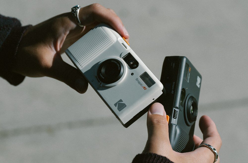Dwie osoby trzymają w rękach aparaty analogowe, jeden biały z logo Kodak, drugi ciemny. Na białym aparacie widoczny jest napis \'Snapic A1\'.