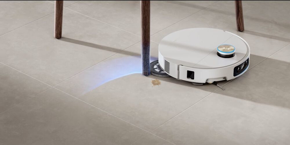 Biały robot sprzątający porusza się po podłodze, omijając nogę stołu. Na podłodze widoczna jest niewielka plama, a robot emituje niebieskie światło.