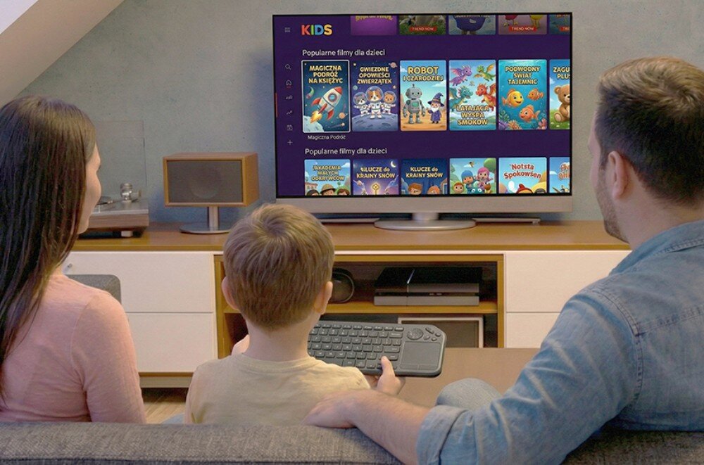 Trzy osoby siedzą na kanapie przed telewizorem, na którym wyświetlane są popularne filmy dla dzieci. Dziecko trzyma klawiaturę z touchpadem.