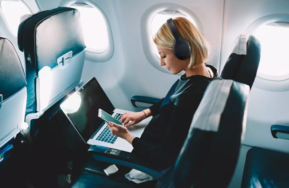 Osoba siedzi w samolocie, używając laptopa i smartfona, mając na głowie czarne słuchawki nauszne. W tle widoczne są okna samolotu i siedzenia pasażerów.