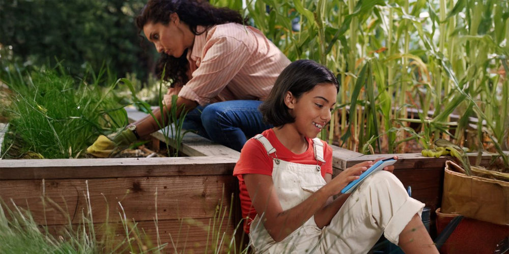 Czytnik e-booków AMAZON Kindle Kids Unicorn Valley 6 cali, Podświetlany ekran, Wi-Fi, Bez reklam Nastolatka siedzi w ogrodzie warzywnym przy drewnianej grządce i korzysta z Kindle Kids, w tle dorosła osoba pielęgnuje rośliny ekran E-Ink z podświetleniem powłoka ograniczająca odblaski regulacja jasności ekran dotykowy przejrzyste menu