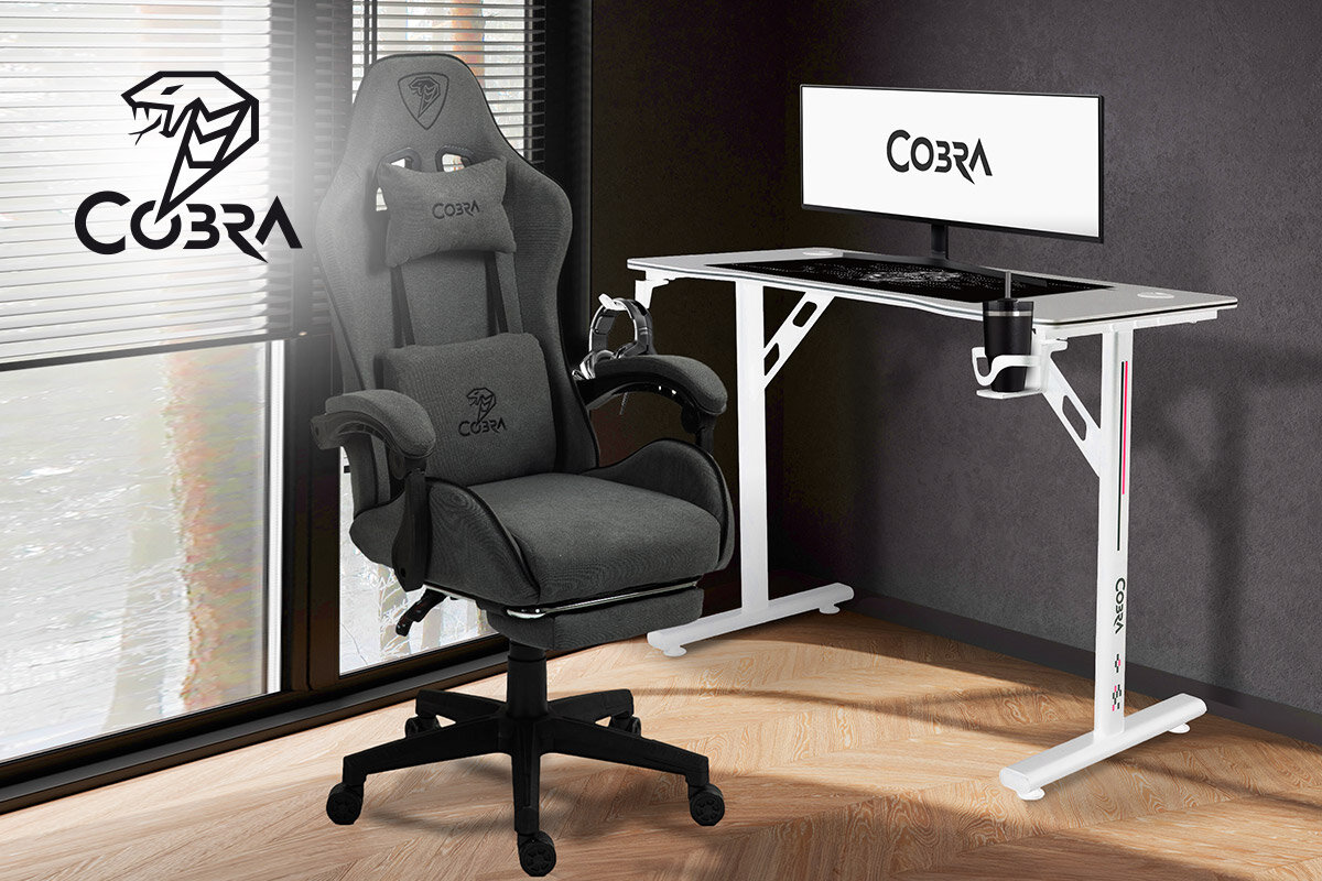 Na obrazie znajduje się biurko gamingowe w kolorze białym z monitorem i klawiaturą oraz szary fotel gamingowy. Na ścianie i monitorze widoczne jest logo COBRA.