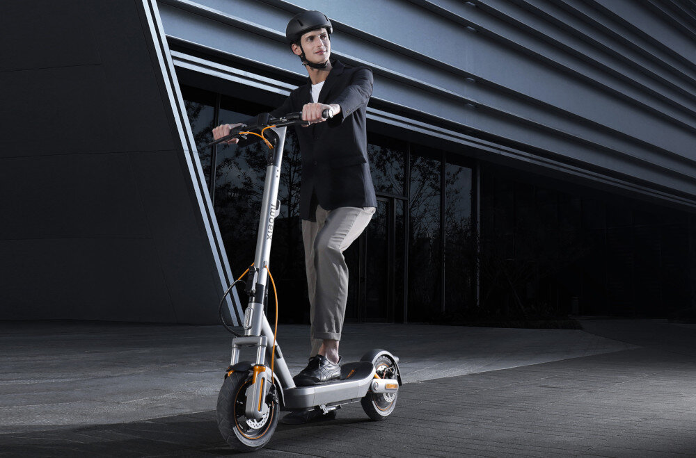 Hulajnoga elektryczna XIAOMI Electric Scooter 6 Max Max 70km 450W 2 amortyzatory 12 cali Czarny miasto ciemny budynek osoba w kasku jedzie na hulajnodze po drodze mobilność środek codziennego transportu wygoda bezpieczeństwo