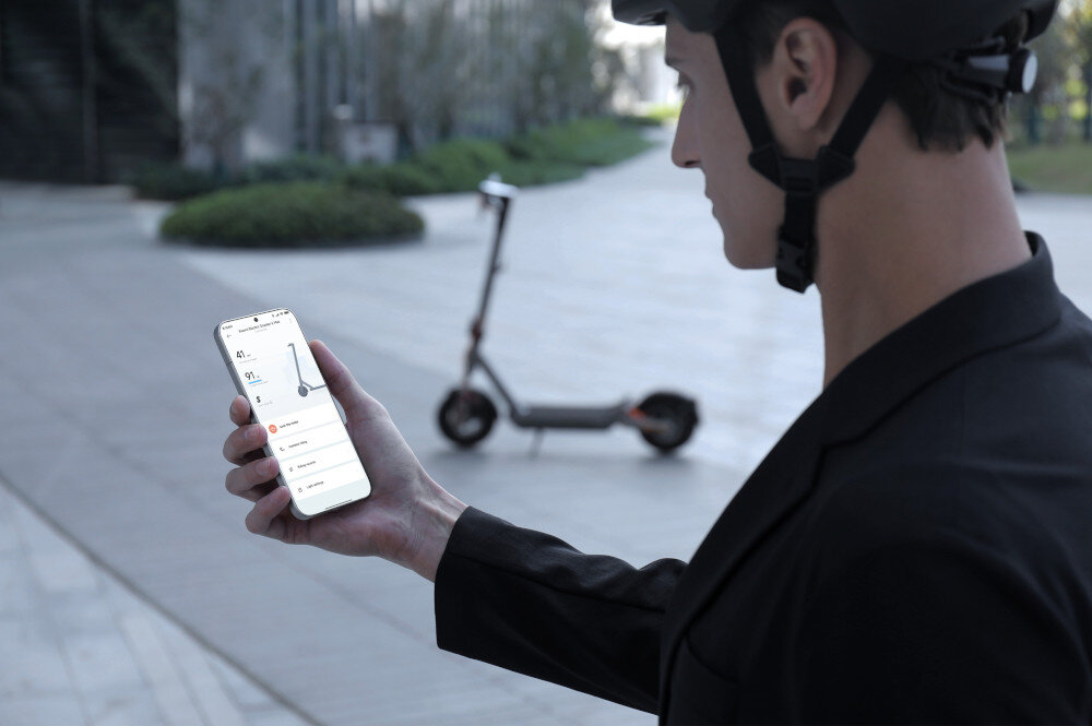 Hulajnoga elektryczna XIAOMI Electric Scooter 6 Max Max 70km 450W 2 amortyzatory 12 cali Czarny miasto osoba z telefonem w dłoni w tle stoi hulajnoga moduł Bluetooth aplikacja XIAOMI Home App na system Android i iOS pełna kontrola nad pojazdem bezpośrednio ze smartfona dodatkowe funkcje diagnostyczne i statystyki jazdy