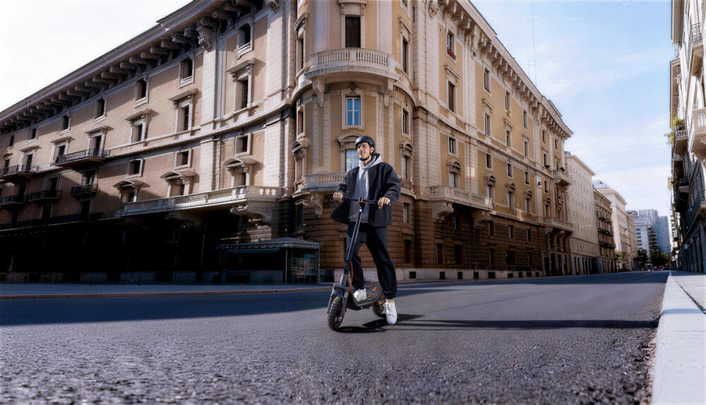 Hulajnoga elektryczna XIAOMI Electric Scooter 6 Pro 70km 400W 2 amortyzatory 12 cali Czarny miasto budynek osoba w kasku opiera się o hulajnogę mobilność środek codziennego transportu wygoda bezpieczeństwo