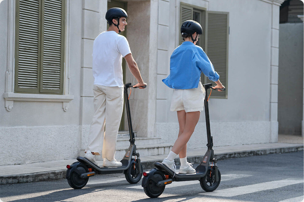 Hulajnoga elektryczna XIAOMI Electric Scooter 6 45km 400W 2 amortyzatory 12 cali Czarny miasto ulica dwie osoby jadą na hulajnogach po ulicy w tle szary budynek z dużymi oknami koła bezdętkowe o średnicy 12 cali niskim oporem toczenia duża przyczepność do podłoża bardzo dobrze absorbują wstrząsy i nierówności terenu opony są odporne na przebicia i nie wymagają konserwacji