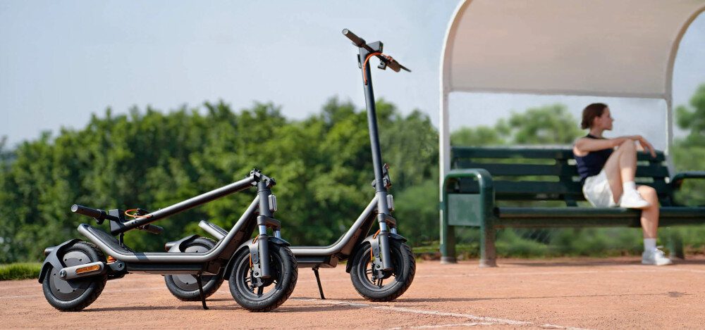 Hulajnoga elektryczna XIAOMI Electric Scooter 6 45km 400W 2 amortyzatory 12 cali Czarny park dwie hulajnogi ustawione na chodniku jedna złożona w tle na ławce siedzi osoba drzewa niebo składana stopka wytrzymała nóżka zamontowana w centralnym punkcie ciężkości sygnał dźwiękowy
