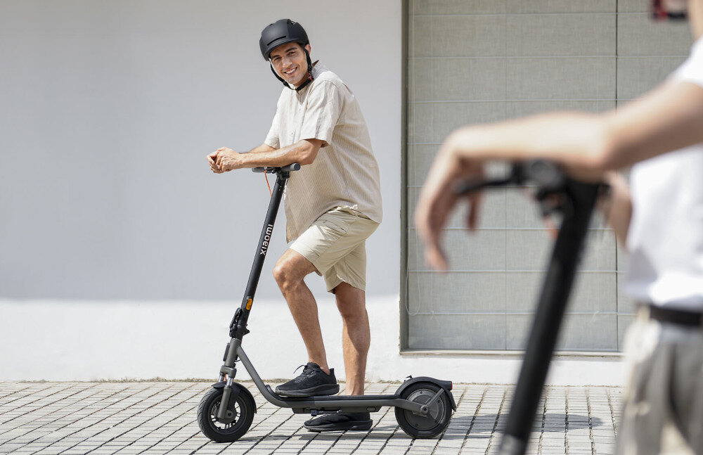 Hulajnoga elektryczna XIAOMI Electric Scooter 6 Lite Lite 25km 300W Amortyzacja 10 cali Czarny miasto ulica osoba opiera się ciałem o hulajnogę patrzy na wprost w tle szary budynek sprawne poruszanie się po mieście wygodne pokonywanie codziennych tras elektryczny napęd pozwala ograniczyć czas dojazdów i zwiększyć niezależność