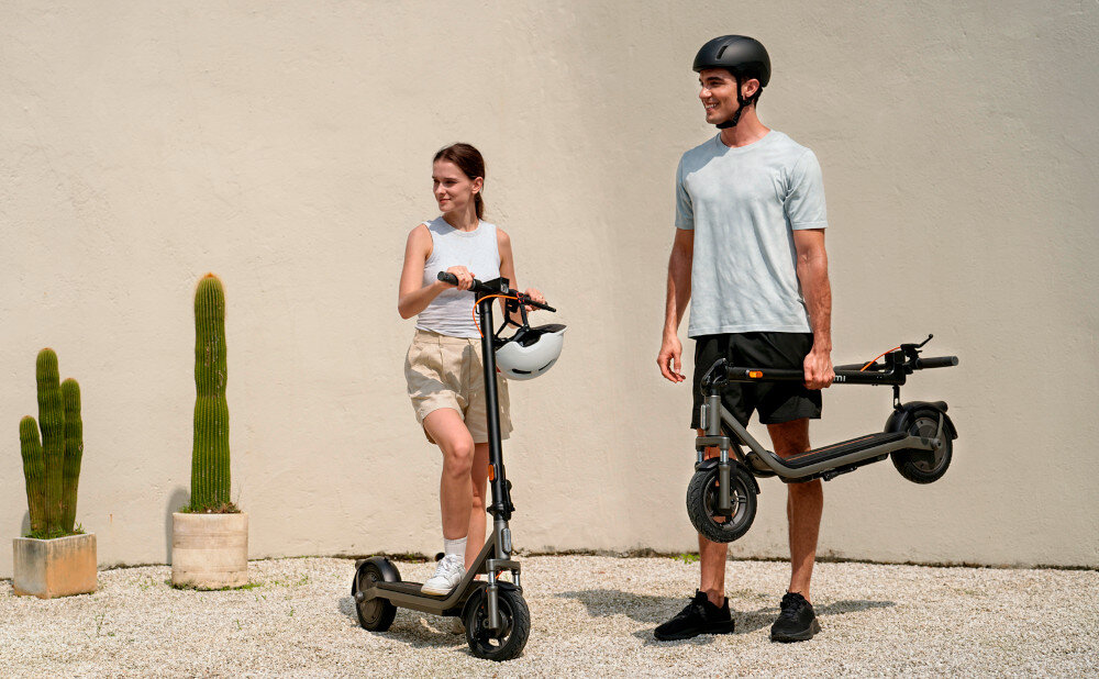 Hulajnoga elektryczna XIAOMI Electric Scooter 6 Lite Lite 25km 300W Amortyzacja 10 cali Czarny jasne tło piaskowy mur dwie osoby jedna osoba trzyma złożoną hulajnogę w dłoni druga opiera się ciałem na hulajnodze obie osoby patrzą w bok składana stopka wytrzymała nóżka zamontowana w centralnym punkcie ciężkości sygnał dźwiękowy