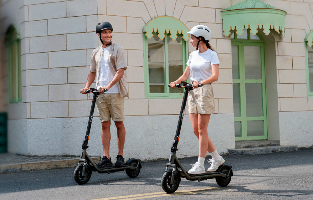 Hulajnoga elektryczna XIAOMI Electric Scooter 6 Lite Lite 25km 300W Amortyzacja 10 cali Czarny miasto dwie osoby jadą na hulajnogach w tle szary budynek z zielonymi drzwiami silnik o mocy 300 W cicha praca i płynna kontrola prędkości pokonuje wzniesienia o nachyleniu do 15% maksymalna prędkość do 20 km/h