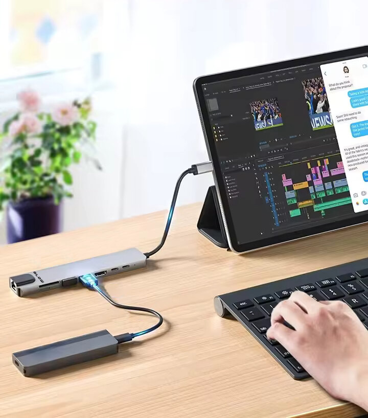 Na drewnianym biurku znajduje się tablet z podłączonym rozdzielaczem USB, obok którego leży klawiatura. Na ekranie tabletu widoczne są aplikacje do edycji wideo i wiadomości tekstowe.
