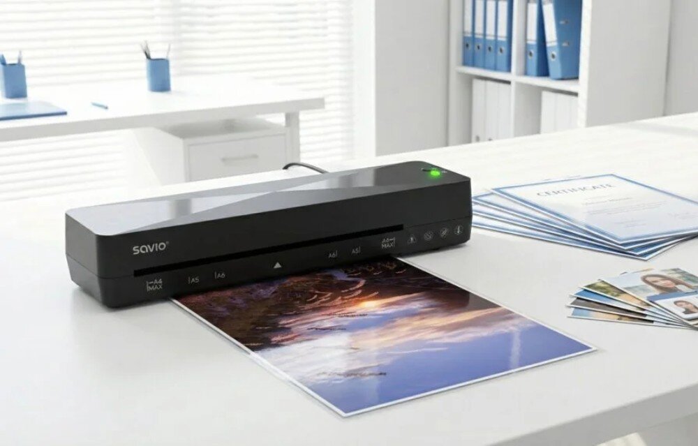 Laminator A4 SAVIO LR-01/B Czarny laminator stoi na białym biurku w biurze i wysuwa zalaminowane kolorowe zdjęcie krajobrazu, a obok leżą gotowe certyfikaty