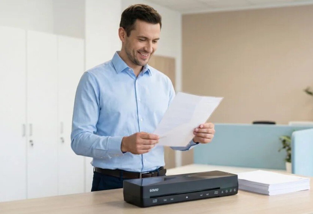 Laminator A4 SAVIO LR-01/B Uśmiechnięty mężczyzna w błękitnej koszuli stoi w biurze i trzyma w dłoniach zalaminowany dokument formatu A4 nad czarnym urządzeniem