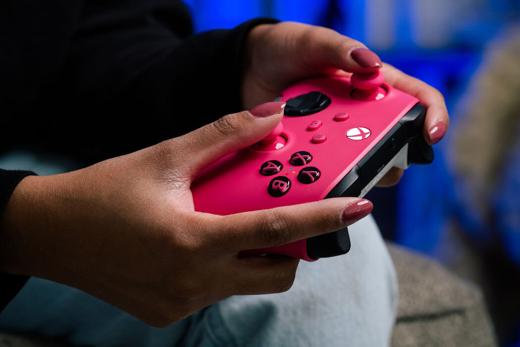 Osoba trzyma różowy kontroler do gier z widocznymi przyciskami i logo Xbox. W tle widoczne są rozmyte niebieskie elementy.