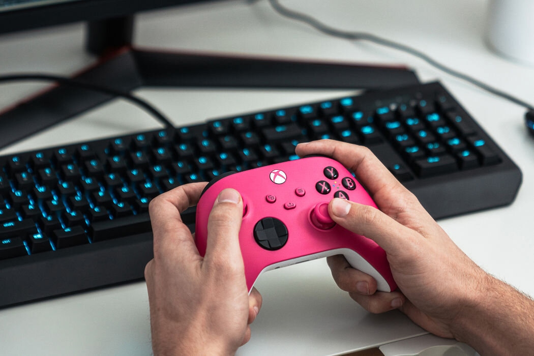 Osoba trzyma różowy kontroler do gier z widocznym logo Xbox. W tle znajduje się klawiatura z podświetlanymi klawiszami.
