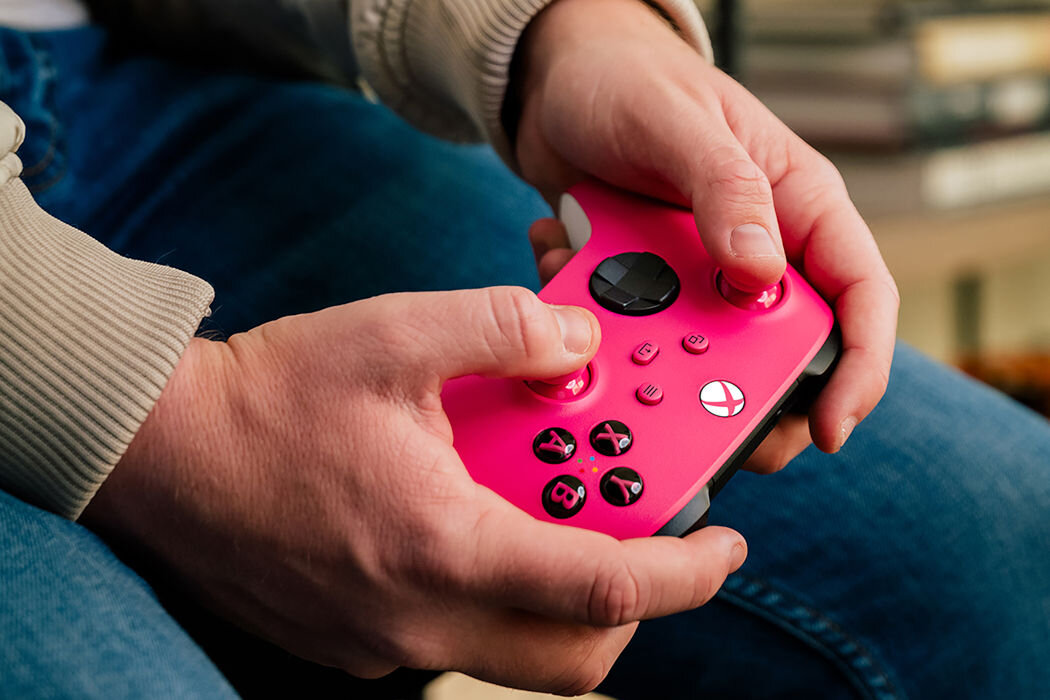 Osoba trzyma różowy kontroler do gier z widocznymi przyciskami i logo Xbox. W tle widać rozmyte elementy wnętrza.