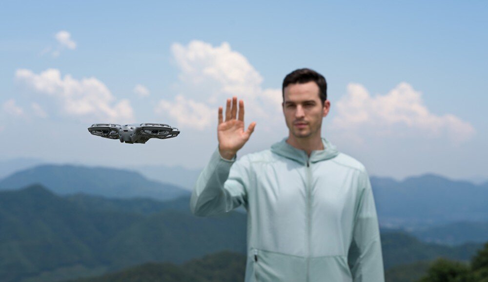 Dron DJI Neo 2 Mężczyzna w jasnej kurtce stoi na górskim tle i unosi dłoń obok małego drona w locie, sterowanie, komendy głosowe, zasięg