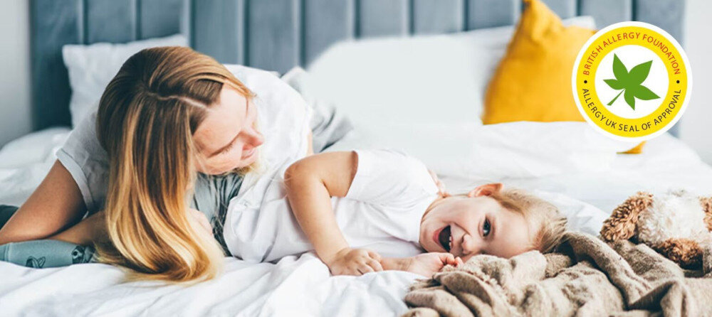 PRALKA LG F4X7511TBB Zdjęcie przedstawia dorosłą osobę z długimi włosami i małe dziecko, które beztrosko leżą razem na białym łóżku. W prawym górnym rogu znajduje się żółte logo British Allergy Foundation z liściem, oznaczające Allergy UK Seal of Approval usuwanie alergenów świeże miękkie pranie dla alergików