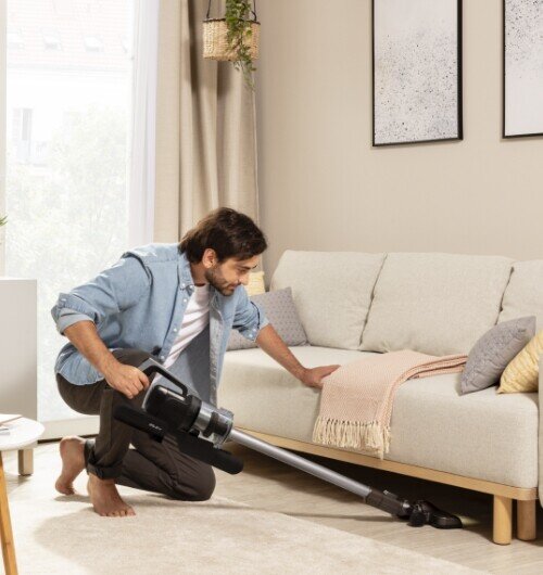 Osoba używa odkurzacza pionowego w salonie, odkurzając przestrzeń pod sofą. W tle widać jasne zasłony i dwa obrazy na ścianie.