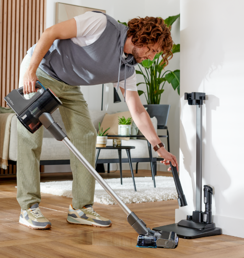 Osoba używa odkurzacza pionowego w jasnym wnętrzu z drewnianą podłogą. W tle widać rośliny doniczkowe i meble.