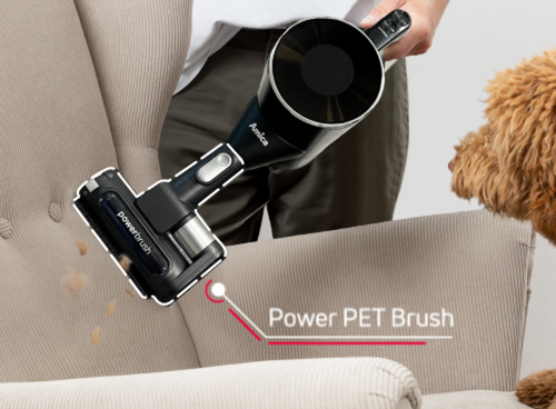 Osoba używa czarnego odkurzacza z napisem \'Amica\' do czyszczenia jasnej tapicerki, obok widoczny jest pies. Na obrazie znajduje się tekst: \'Power PET Brush\'.