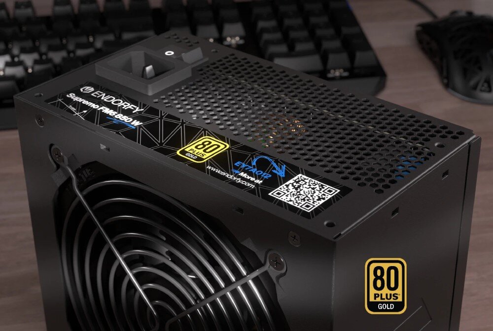 Komputer MAD DOG ENDORFY500ARGB-I25WR32WH Zbliżenie na zasilacz ustawiony na biurku. Widoczna jest naklejka informacyjna z oznaczeniem modelu, certyfikatem 80 PLUS Gold, kodem QR. W tle widać fragment klawiatury i myszy komputerowej, zasilacz, moc, sprawność, kondensatory