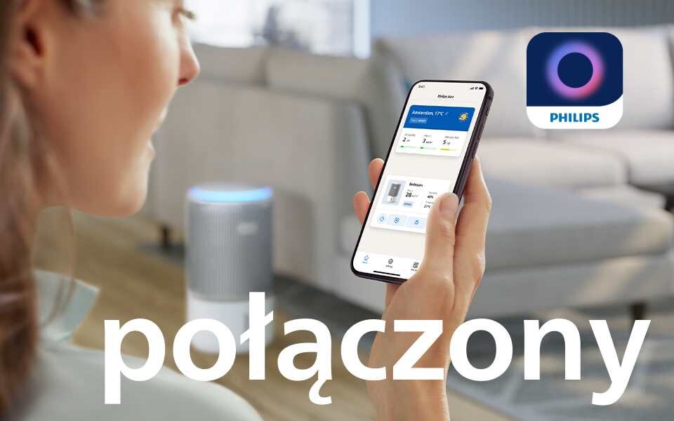 Osoba trzyma smartfon z aplikacją pokazującą dane dotyczące jakości powietrza. W tle widoczny jest oczyszczacz powietrza, a na obrazie znajduje się napis \'połączony\' oraz logo PHILIPS.