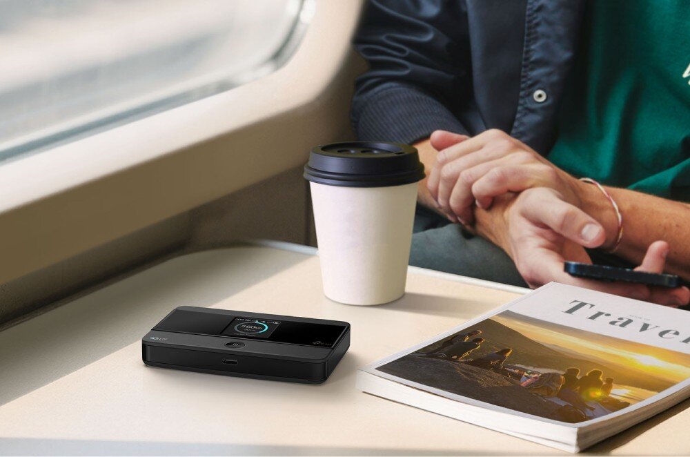 Router TP-LINK M7352 Czarny, prostokątny router mobilny z małym ekranem leżący na jasnym stoliku w pociągu obok kubka kawy i książki