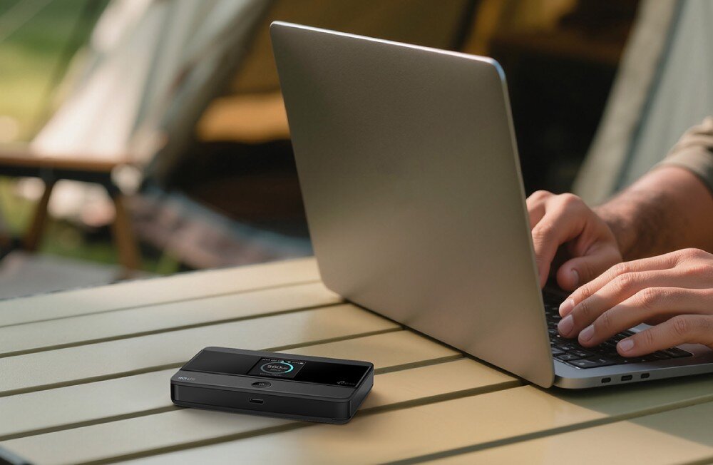 Router TP-LINK M7352 Mały router bezprzewodowy umieszczony na drewnianym stole kempingowym przed otwartym laptopem