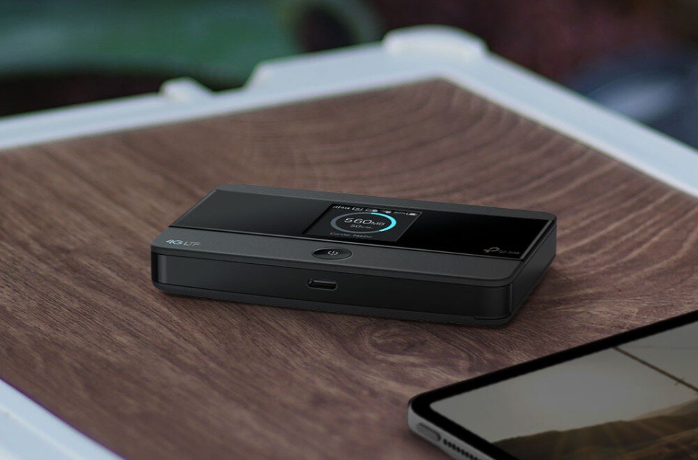 Router TP-LINK M7352 Zbliżenie na czarny router mobilny z włączonym wyświetlaczem, ustawiony na drewnianej powierzchni obok krawędzi tabletu