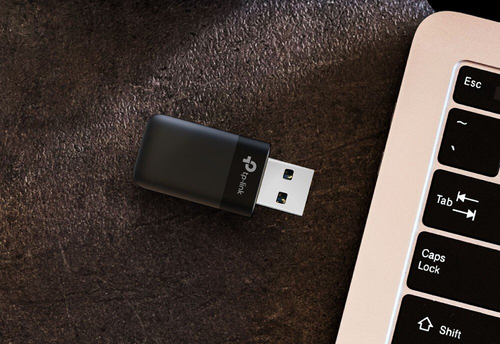 Karta sieciowa TP-LINK Archer TX23U Mały czarny adapter USB leżący na ciemnym blacie obok krawędzi srebrnego laptopa