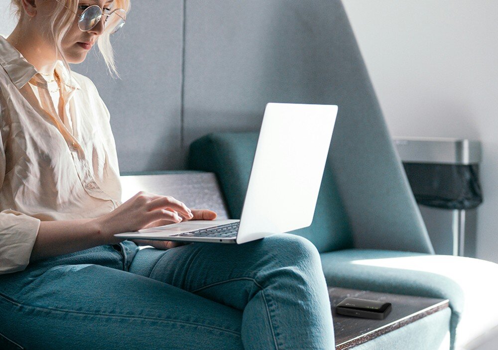 Router TP-LINK M7005 Kobieta w okularach siedząca na turkusowej kanapie z laptopem na kolanach, obok którego na stoliku leży mały czarny router