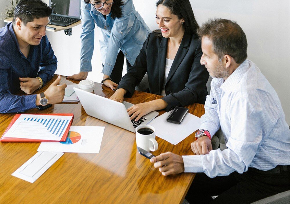 Router TP-LINK M7005 Grupa czterech osób siedząca wokół drewnianego stołu konferencyjnego, na którym obok otwartego laptopa leży czarny router mobilny