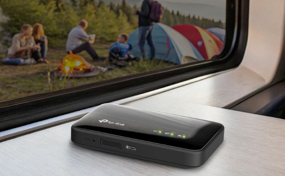 Router TP-LINK M7005 Czarny router mobilny leżący na jasnej powierzchni parapepu lub stolika, z widokiem przez okno na grupę ludzi siedzącą przy ognisku obok namiotów